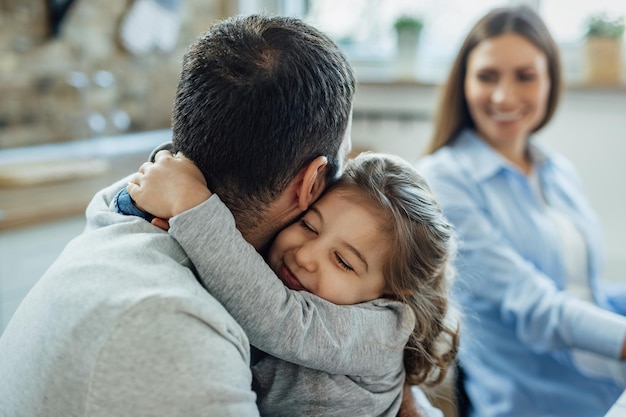 Niña cariñosa con los ojos cerrados abrazando a su padre en casa La madre está en el fondo