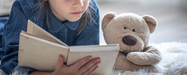 Una niña en casa, tirada en el suelo con su juguete favorito y lee un libro.