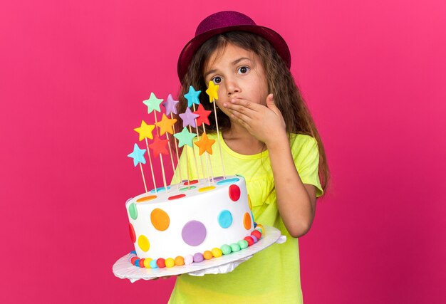 Niña caucásica ansiosa con gorro de fiesta púrpura poniendo la mano en la boca y sosteniendo el pastel de cumpleaños aislado en la pared rosa con espacio de copia