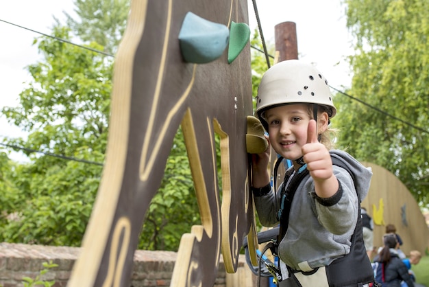 Foto gratuita niña caucásica con un casco blanco y equipo de parque de aventuras sonriendo y sosteniendo el pulgar hacia arriba