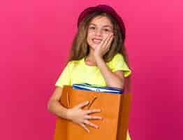 Foto gratuita niña caucásica complacida con gorro de fiesta púrpura poniendo la mano en la cara y sosteniendo la caja de regalo en una bolsa de papel aislada en la pared rosa con espacio de copia