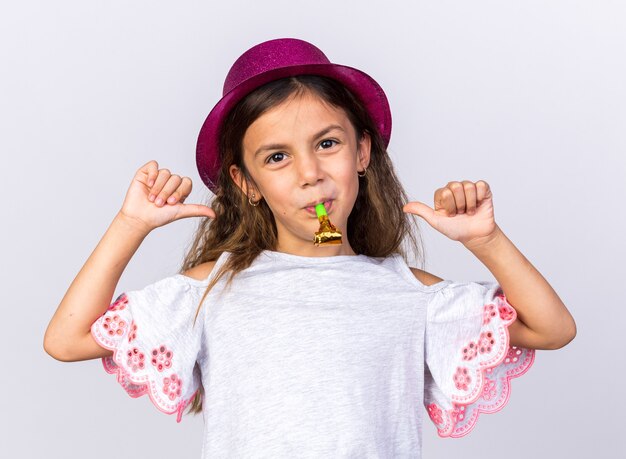 Foto gratuita niña caucásica complacida con gorro de fiesta púrpura soplando un silbato de fiesta y apuntando a sí misma aislada en la pared blanca con espacio de copia