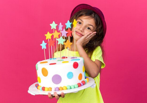 Niña caucásica complacida con gorro de fiesta púrpura sosteniendo pastel de cumpleaños y poniendo la mano en la cara aislada en la pared rosa con espacio de copia