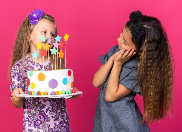 Niña caucásica complacida mirando a la niña rubia sosteniendo pastel de cumpleaños aislado en la pared rosa con espacio de copia