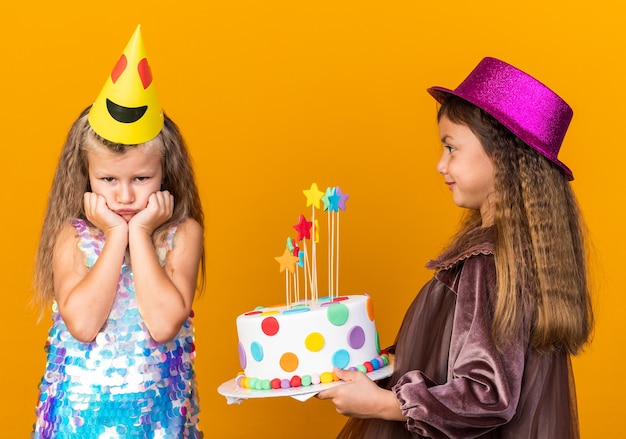 Niña caucásica complacida con sombrero de fiesta púrpura sosteniendo pastel de cumpleaños y mirando triste niña rubia con gorra de fiesta aislada en pared naranja con espacio de copia