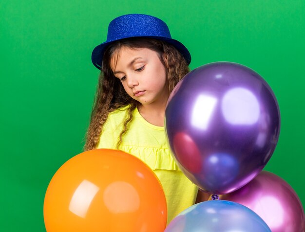 Niña caucásica decepcionada con gorro de fiesta azul sosteniendo globos de helio y mirando hacia abajo aislado en la pared verde con espacio de copia