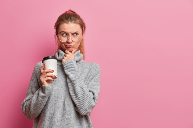 Niña caucásica disgustada piensa profundamente con expresión infeliz, toca la barbilla y frunce los labios, piensa cómo resolver una situación problemática, usa un suéter gris