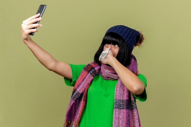 Niña caucásica enferma con gorro de invierno y bufanda tomando selfie sosteniendo la servilleta manteniendo la mano en la boca aislada sobre fondo verde oliva con espacio de copia