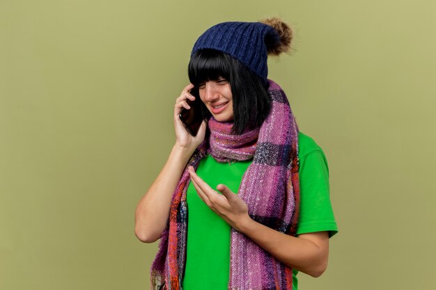 Niña caucásica enferma joven disgustada con gorro de invierno y bufanda hablando por teléfono mirando hacia abajo mostrando la mano vacía aislada sobre fondo verde oliva con espacio de copia