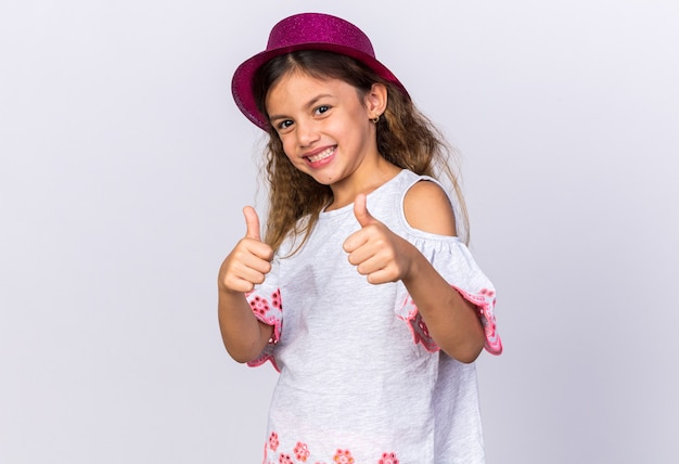 Foto gratuita niña caucásica ittle sonriente con gorro de fiesta púrpura thumbs up aislado en una pared blanca con espacio de copia