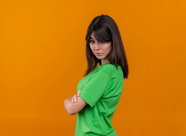 Niña caucásica joven sorprendida en camisa verde se encuentra de lado con los brazos cruzados sobre fondo naranja aislado con espacio de copia