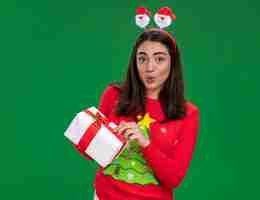 Foto gratuita niña caucásica joven sorprendida con diadema de santa tiene caja de regalo de navidad aislada en la pared verde con espacio de copia