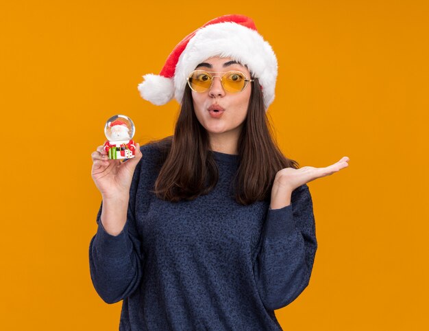 Niña caucásica joven sorprendida en gafas de sol con gorro de Papá Noel sostiene el globo de nieve y mantiene la mano abierta aislada en la pared naranja con espacio de copia