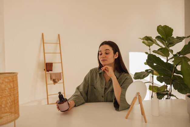 Niña caucásica lee cuidadosamente la composición de la crema de manos en el frasco mientras se sienta a la mesa en el interior Morena con camisa verde convencida de la naturalidad del producto Concepto de cuidado