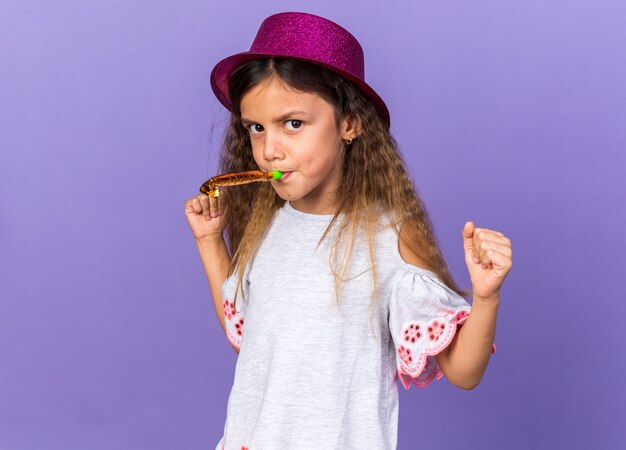 Niña caucásica molesta con gorro de fiesta violeta soplando el silbato de fiesta y manteniendo los puños arriba aislados en la pared púrpura con espacio de copia