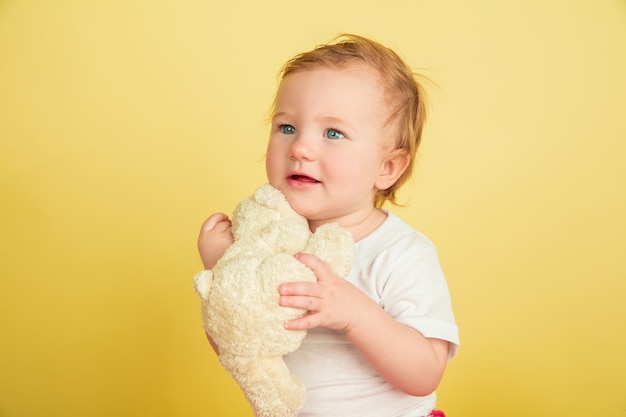 Niña caucásica, niños aislados sobre fondo amarillo de estudio. Retrato de niño lindo y adorable, bebé jugando con osito de peluche. Concepto de infancia, familia, felicidad, nueva vida, educación.