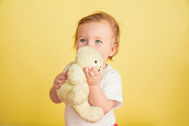 Niña caucásica, niños aislados sobre fondo amarillo de estudio. Retrato de niño lindo y adorable, bebé jugando con osito de peluche.