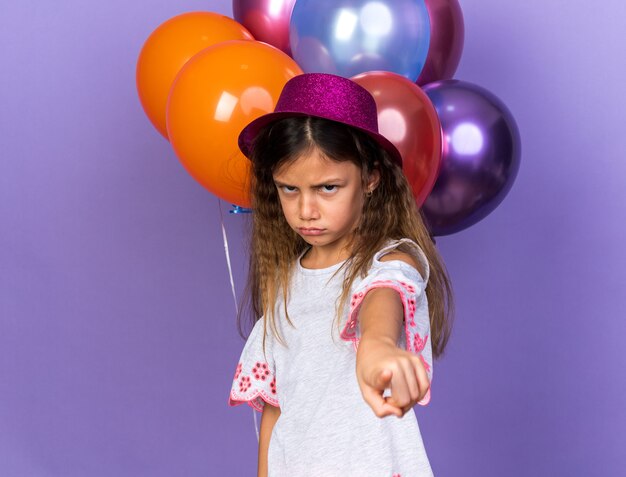 Niña caucásica ofendida con gorro de fiesta violeta apuntando de pie delante de globos de helio aislado en la pared púrpura con espacio de copia