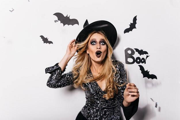 Niña caucásica soñadora con sombrero de mago expresando emociones de sorpresa. Foto interior de mujer europea feliz en traje de bruja celebrando halloween.