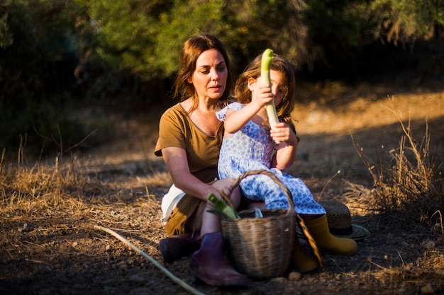 Niña de la celebración de verduras sentado en el regazo de su madre en el campo