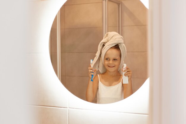 Niña cepillándose los dientes en el baño, mirando su reflejo en el espejo con expresión facial positiva y sonrisa, vistiendo una camiseta blanca y envuelto el pelo en una toalla.