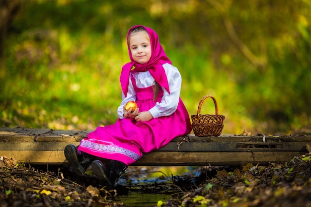 Niña con un chal rosa y vestido como Masha y el oso de la caricatura sostiene una canasta de mimbre y recoge manzanas.