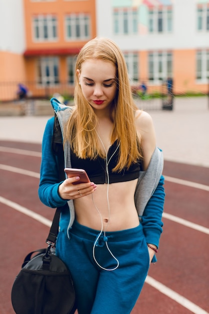 Una niña con un chándal azul y una camiseta corta va al estadio. Lleva una bolsa. Ella está escuchando la música.