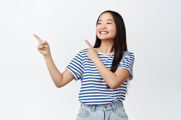 Niña china feliz señalando con el dedo y mirando a la izquierda en el anuncio sonriendo complacida mostrando la promoción o el logotipo de la tienda de pie sobre fondo blanco.