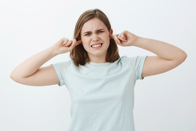 La niña cierra los oídos sintiéndose disgustada cuando la gente pelea cerca de ella. Intensa mujer joven insatisfecha apretando los dientes por la incomodidad sintiéndose molesto por el sonido fuerte que cubre la audición con tapones para los oídos