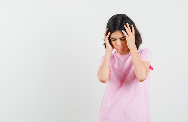 Niña cogidos de la mano a la cabeza en camiseta rosa y con aspecto cansado. vista frontal.