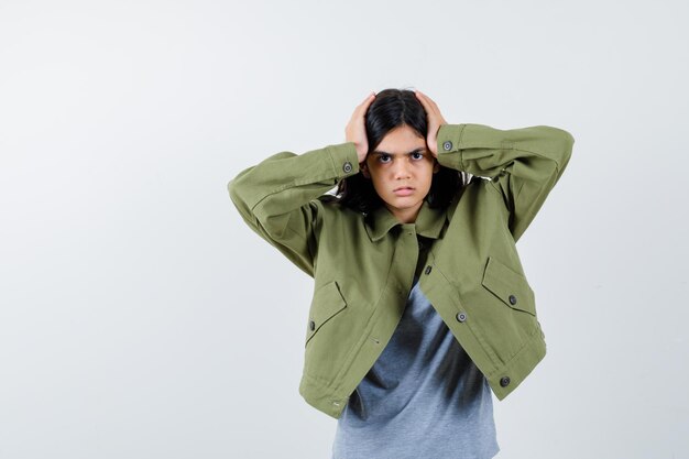 Niña cogidos de la mano en la cabeza con suéter gris, chaqueta caqui, pantalón de mezclilla y mirando serio, vista frontal.