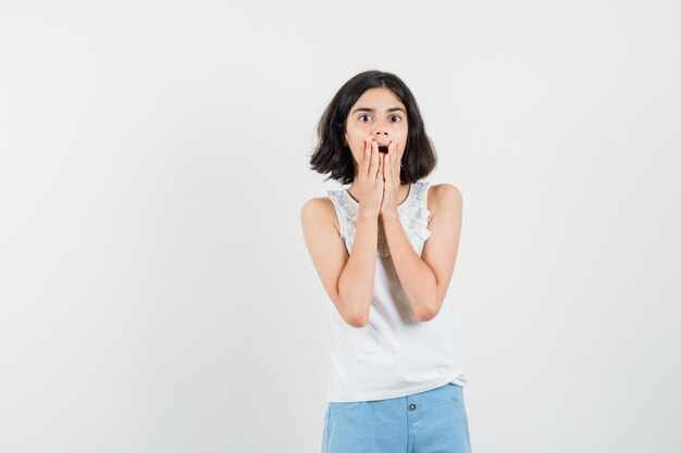 Niña cogidos de la mano cerca de la boca en blusa blanca, pantalones cortos y mirando sorprendida. vista frontal.