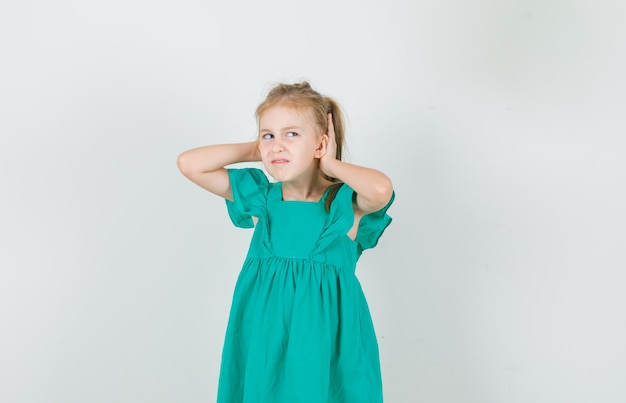 Niña cogidos de la mano detrás de las orejas en vestido verde y mirando disgustado. vista frontal.