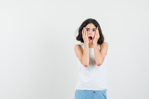 Niña cogidos de la mano en las mejillas en blusa blanca, pantalones cortos y mirando sorprendido, vista frontal.