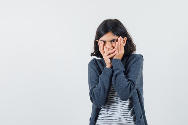 Niña cogidos de la mano en las mejillas en camiseta, chaqueta y mirando triste