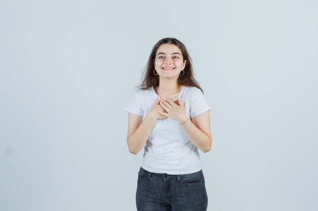 Niña cogidos de la mano en el pecho en camiseta, jeans y mirando feliz. vista frontal.