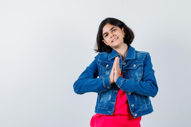 Foto gratuita niña cogidos de la mano en posición de oración en camiseta roja y chaqueta de jean y mirando alegre. vista frontal.