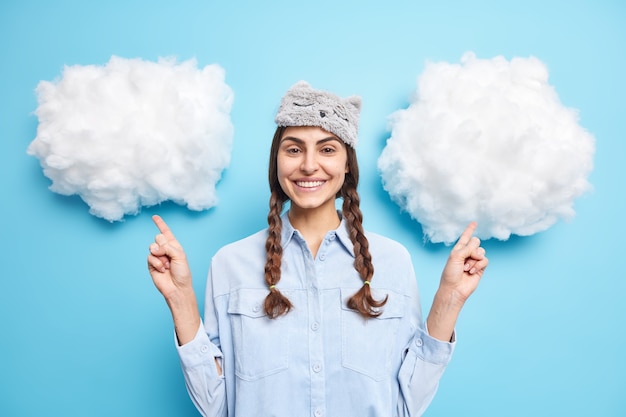 Foto gratuita niña con coletas lleva antifaz y camisa casual puntos arriba en nubes blancas sonríe suavemente producto demostrado para dormir aislado en azul