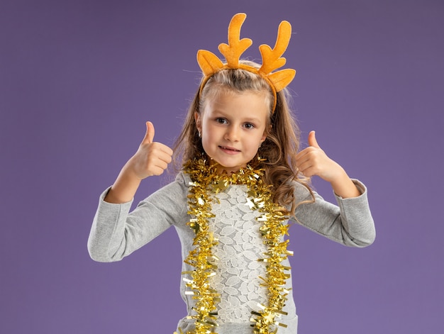 Niña complacida con aro de pelo de navidad con guirnalda en el cuello mostrando los pulgares hacia arriba aislado sobre fondo azul.