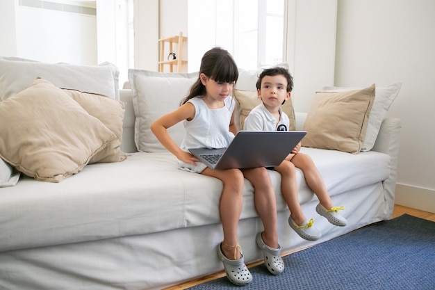 Niña concentrada y su hermano pequeño sentados en el sofá en casa, usando la computadora portátil para videollamadas, chat en línea, viendo videos o películas.