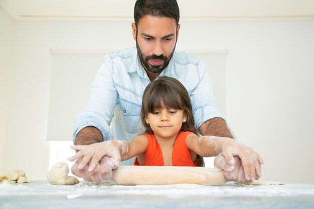 Niña concentrada y su papá amasando y rodando masa sobre la mesa de la cocina con harina desordenada.