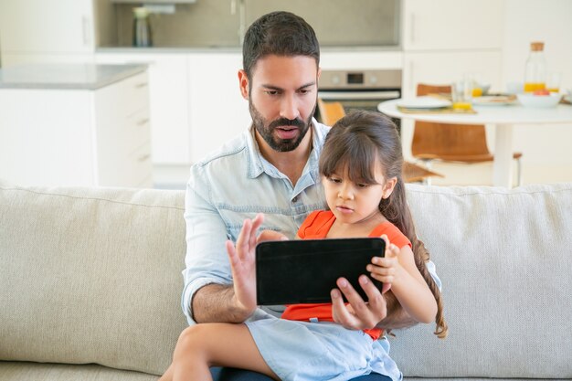 Niña concentrada y su papá usando la aplicación en línea, viendo películas o leyendo juntos en la pantalla de la tableta.