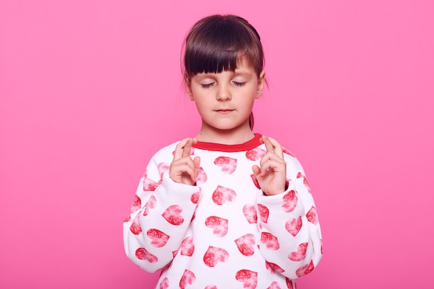 niña concentrada vistiendo un jersey casual con corazones manteniendo los ojos cerrados y los dedos cruzados, aislado sobre una pared rosa.