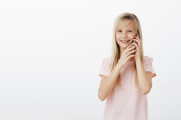 Niña en contacto con abuelos que viven lejos. Retrato de niño adorable complacido en camiseta rosa, hablando por teléfono inteligente y sosteniendo el dispositivo con ambas manos, sonriendo ampliamente