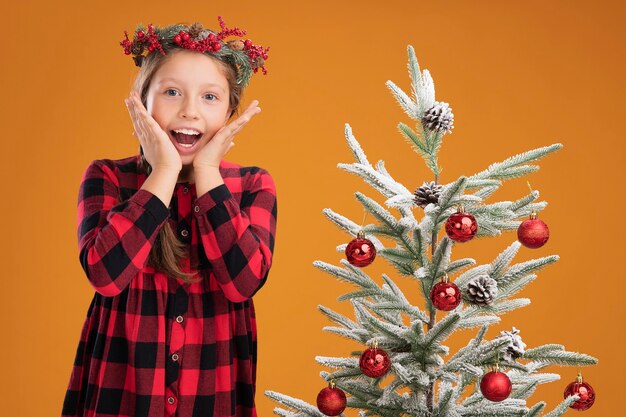 Niña con corona de navidad en camisa a cuadros feliz y emocionado de pie junto a un árbol de navidad sobre la pared naranja