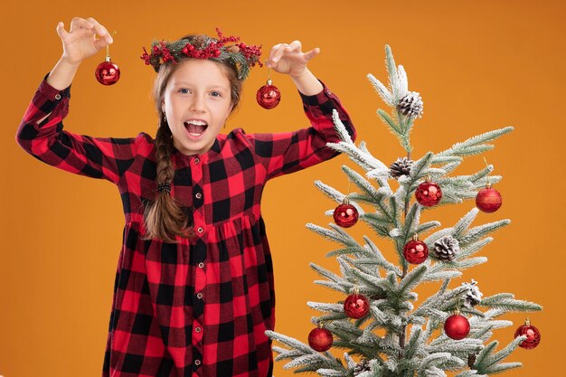 Niña con corona de Navidad en camisa a cuadros sosteniendo bolas de Navidad mirando a la cámara feliz y sonriente de pie junto a un árbol de Navidad sobre fondo naranja