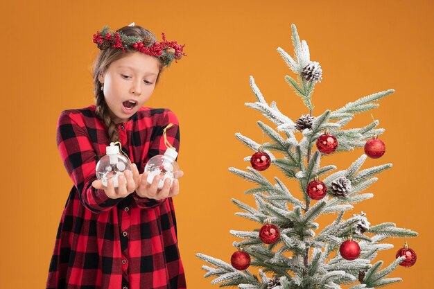 Niña con corona de navidad en camisa a cuadros sosteniendo bolas de navidad mirándolos confundido y sorprendido de pie junto a un árbol de navidad sobre una pared naranja