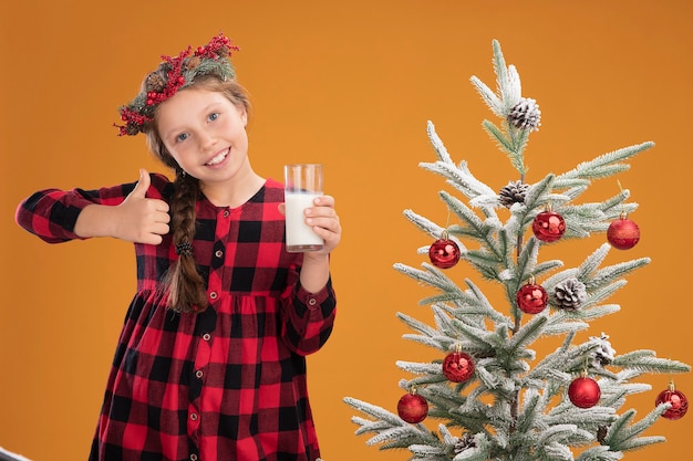 Niña con corona de Navidad en camisa a cuadros sosteniendo un vaso de leche sonriendo alegremente mostrando los pulgares para arriba de pie junto a un árbol de Navidad sobre la pared naranja