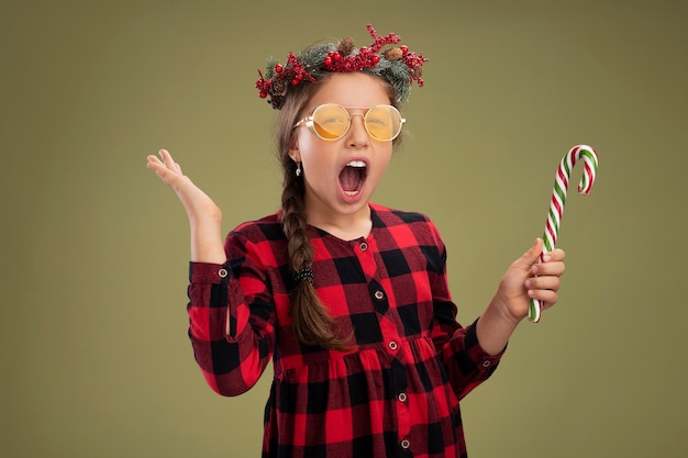 Niña con corona de Navidad en vestido comprobado sosteniendo bastón de caramelo mirando a la cámara feliz y emocionado gritando levantando el brazo sobre fondo verde