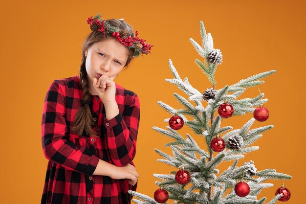 Niña con corona de navidad en vestido de cuadros con la mano en la barbilla pensando con cara seria de pie junto a un árbol de navidad sobre la pared naranja